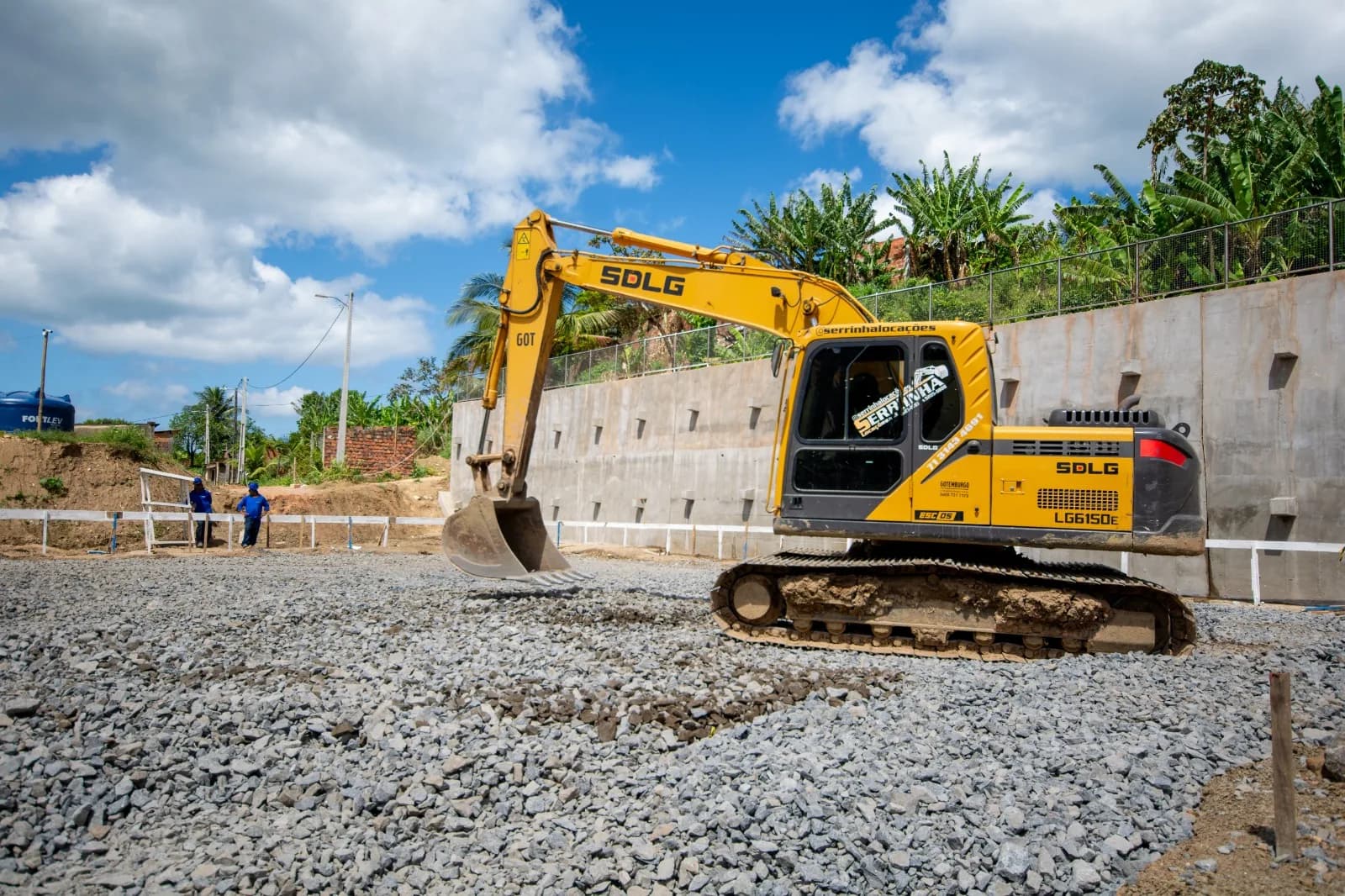 Muro de arrimo em concreto armado com barbacãs de drenagem, com escavadeira sobre plataforma de brita em primeiro plano