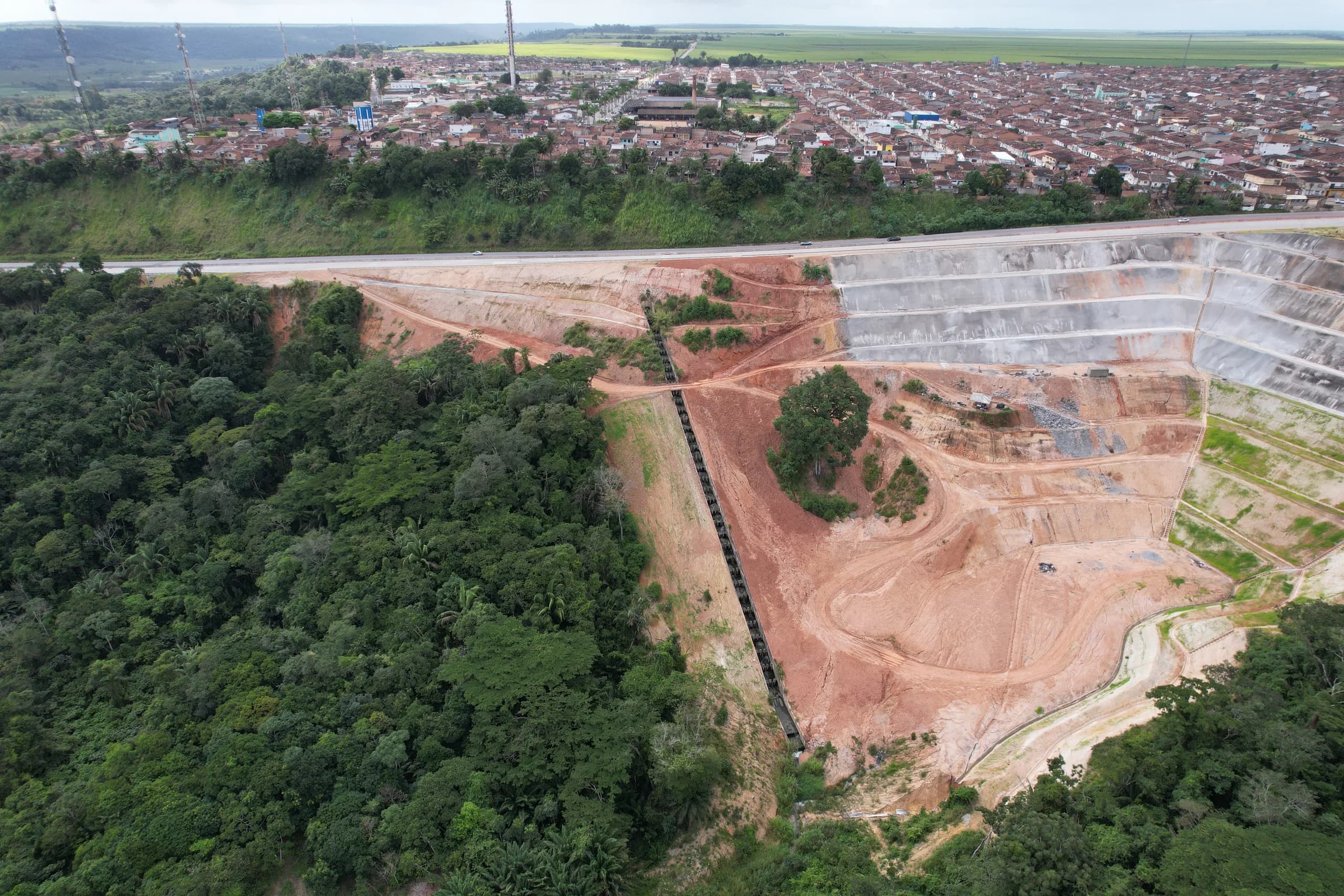 Vista aérea de obra de contenção de encosta às margens de rodovia, com taludes escalonados revestidos em concreto projetado e área urbana ao fundo